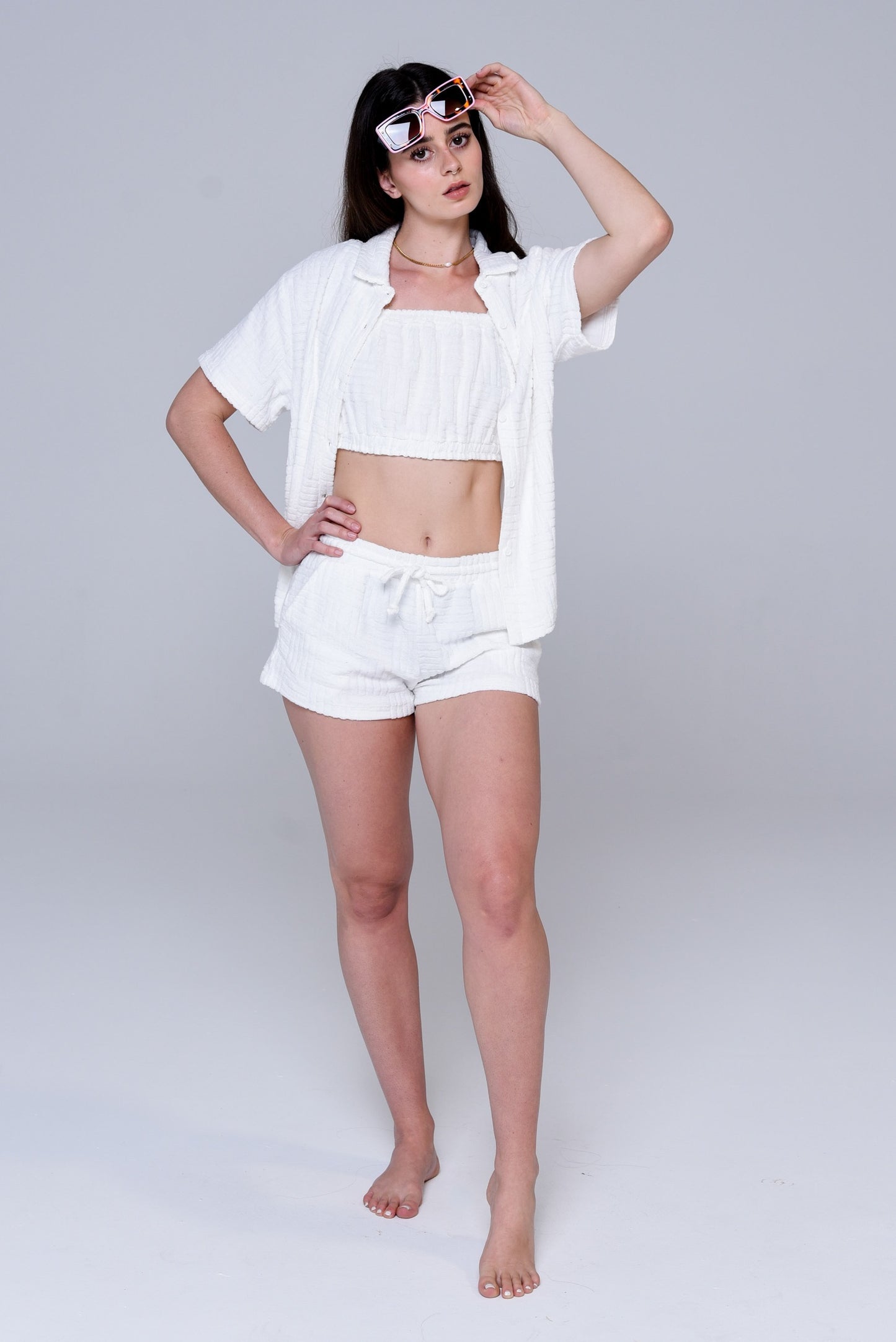 Brunette wearing a white textured terry set of a button front shirt, tube top and short shorts. She has sun glasses on her forehead. 