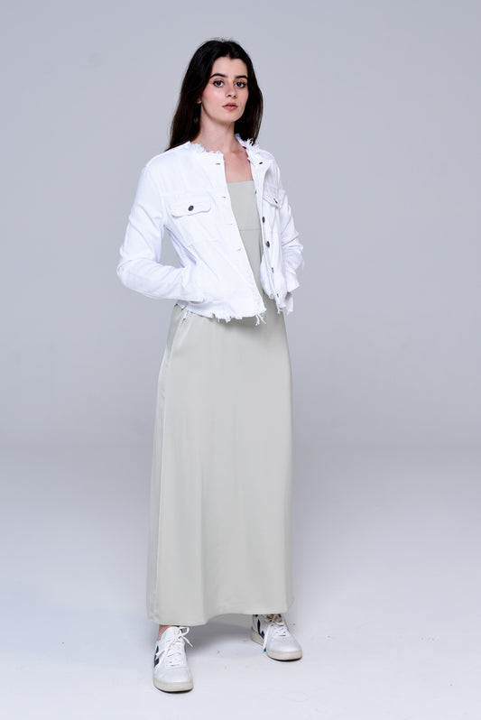 Brunette wearing a white denim collarless jacket with a tan maxi slip dress and white sneaker.