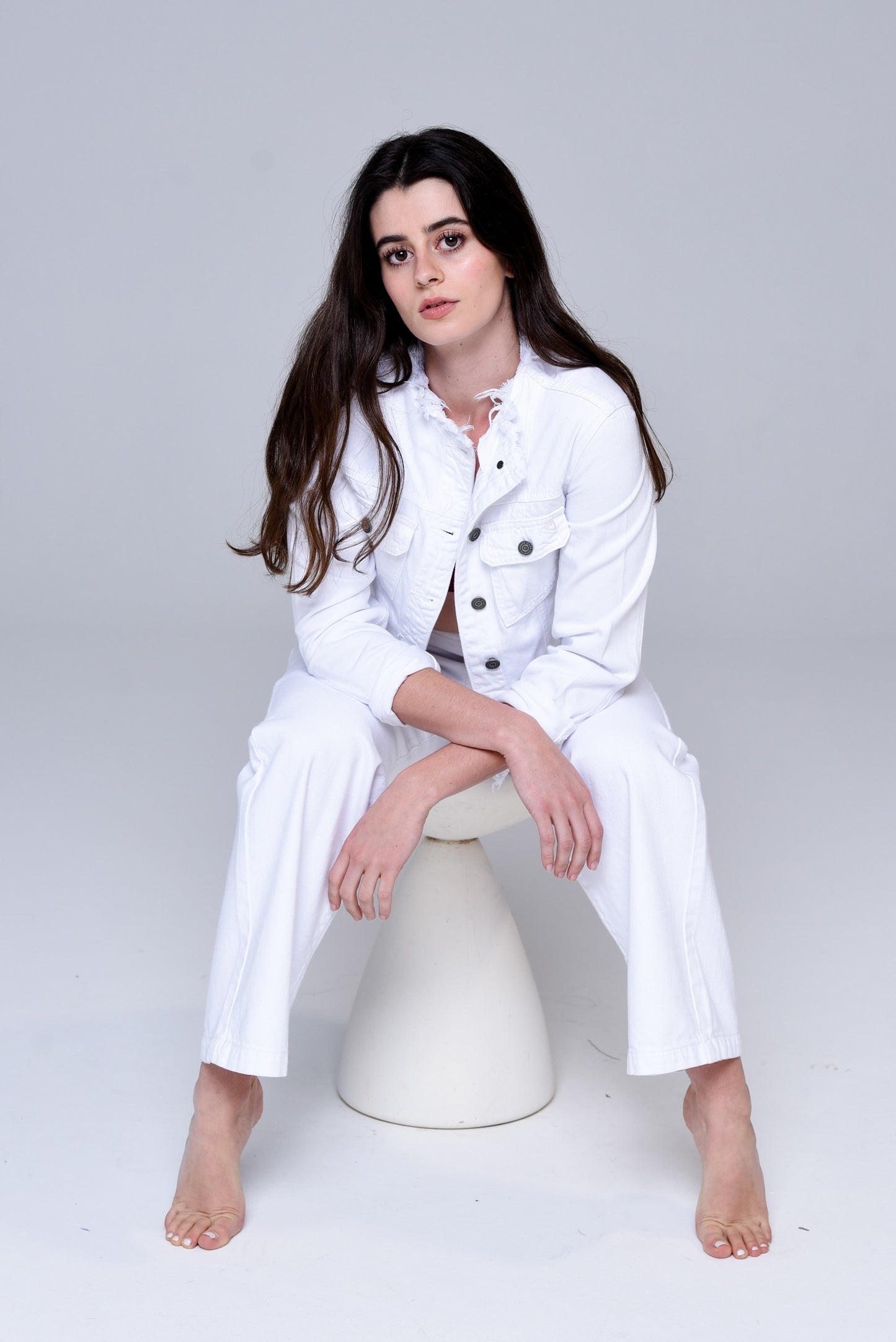 Brunette sitting on a stool wearing a white denim collarless jacket, burgundy bra and white jeans.